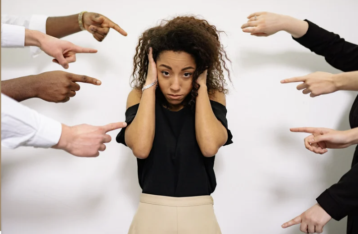 girl in black shirt, standing with her hands over her ears. Multiple arms on the sides are pointing at her
